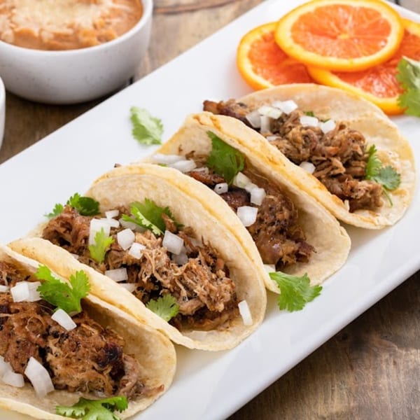 Four El Tequila Mexican Bar & Grill carnitas tacos with cilantro and diced onions on a white plate.